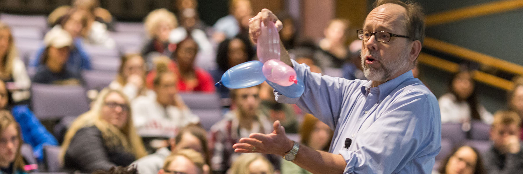 Professor holding balloons for chemistry experiment in front of auditorium with students